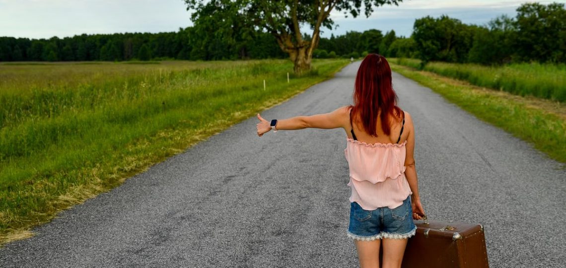 Woman hitchhiking on a rural road in Vihasoo, Estonia, carrying vintage luggage.